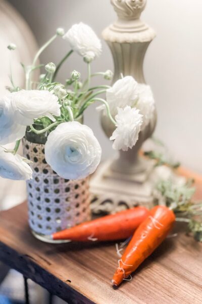 A UPDATED GLASS VASE WITH CANE FULL OF WHITE FLOWERS AND TWO FAUX CARROTS LAYING BESIDE.