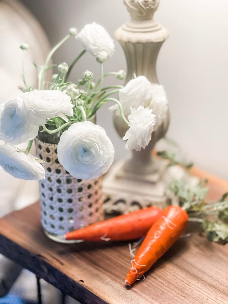 A UPDATED GLASS VASE WITH CANE FULL OF WHITE FLOWERS AND TWO FAUX CARROTS LAYING BESIDE.